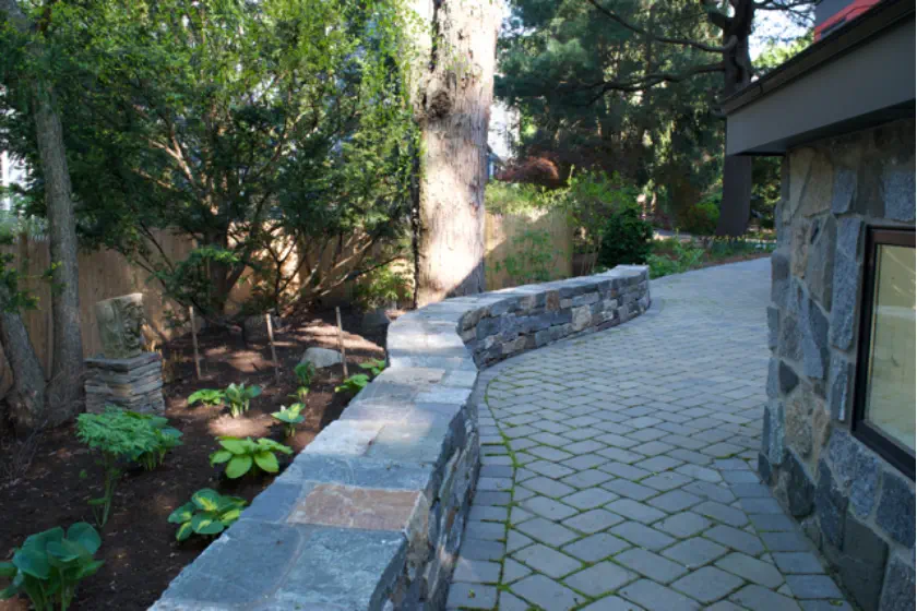 A curved stone walkway bordered by a stone wall runs alongside a garden with leafy plants and tall trees, next to a building with a stone exterior. Sunlight filters through the trees, casting shadows on the ground.