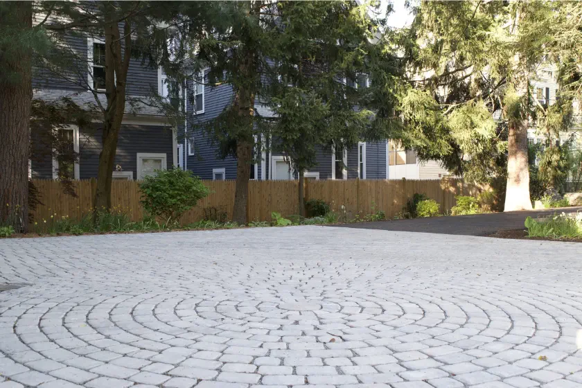 A circular pattern of white paving stones forms a driveway in front of a wooden fence, with tall trees and blue houses in the background on a sunny day.
