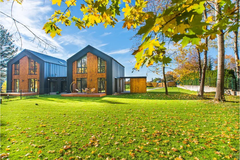 Modern house with large windows and dark roof sits on a spacious green lawn, scattered with autumn leaves. Trees with yellow foliage frame the bright, sunny outdoor scene.