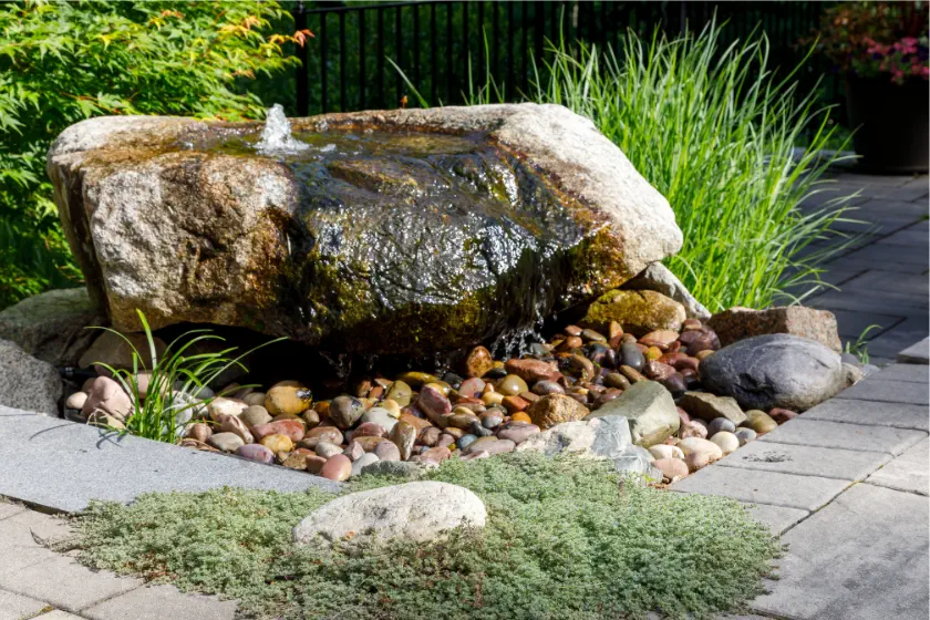A small garden fountain features water flowing over a large rock onto smooth river stones, surrounded by green plants and bordered by stone pavers. Sunlight highlights the natural textures.