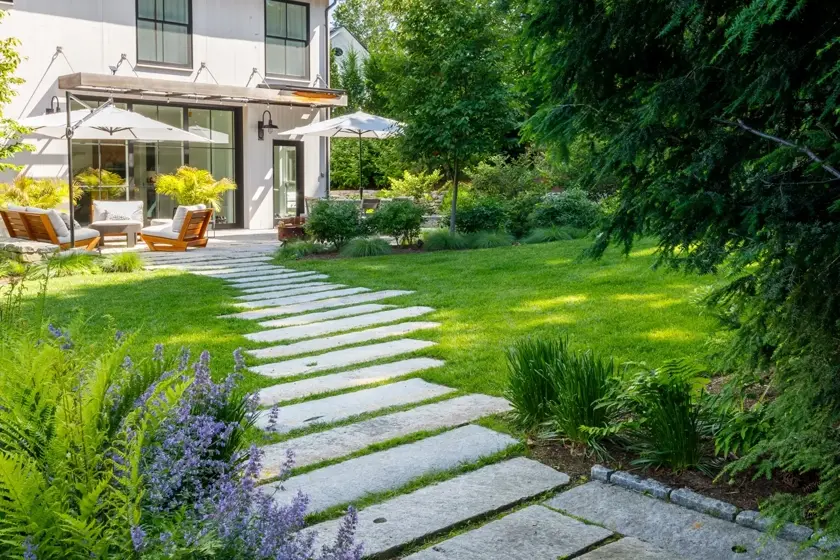 Rectangular stone pavers form a winding path through a lush green garden to a modern house with patio seating, large umbrellas, and plants, surrounded by trees and flowering bushes.