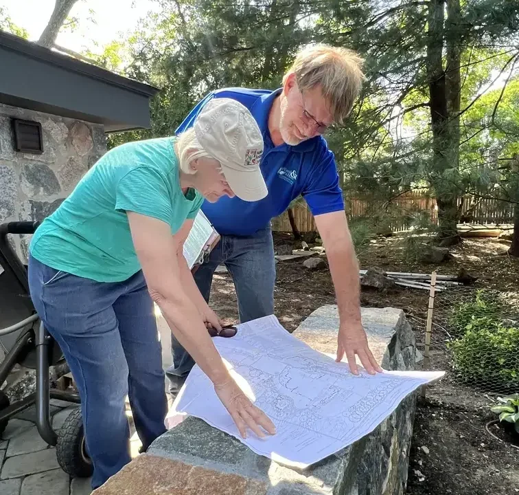Two people stand outdoors by a stone wall, looking at a large map or blueprint spread out in front of them. Trees, greenery, and garden tools are visible in the background.