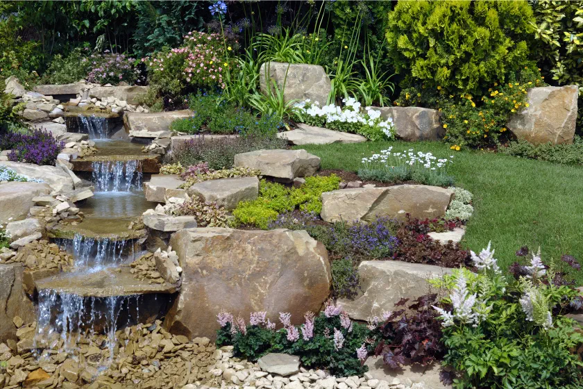 A landscaped garden with a small cascading waterfall flowing over rocks, surrounded by green grass, colorful flowers, shrubs, and large stones, creating a peaceful, natural scene.