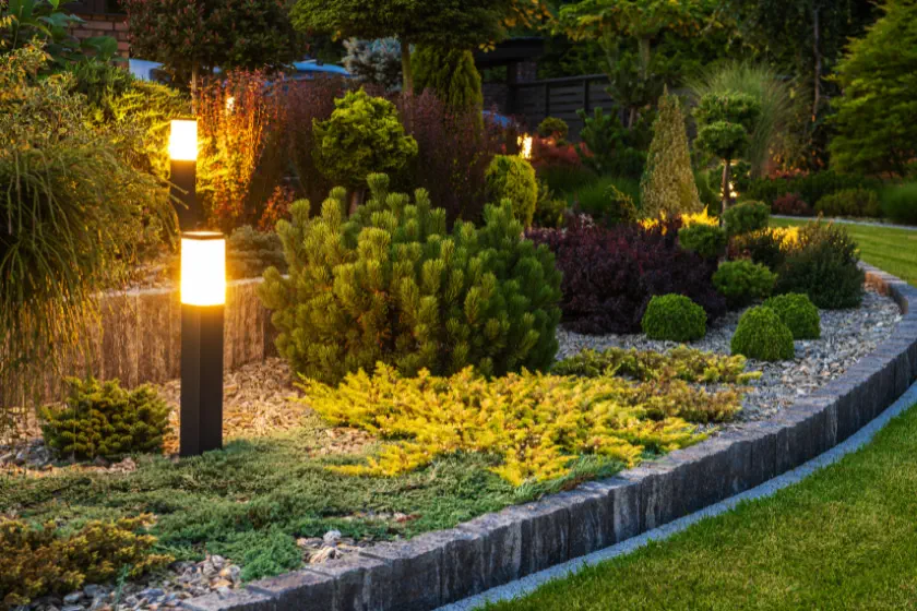 Garden Enhancement 3 A well-maintained garden bed with various bushes, shrubs, and groundcover plants, bordered by stone edging and illuminated by modern outdoor lights at dusk.