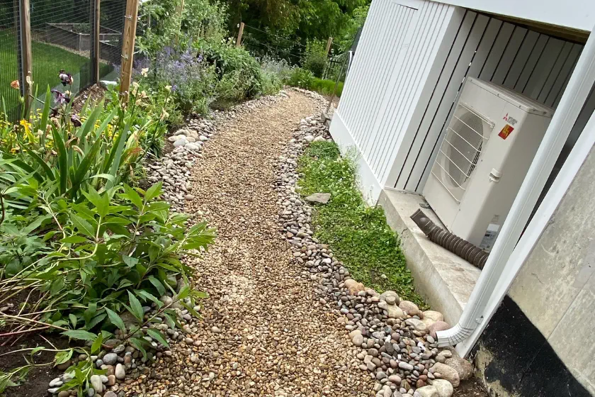 A gravel path winds beside a building with an air conditioning unit, bordered by green plants and flowers on one side and river rocks along the edges. A fenced garden area is visible in the background.