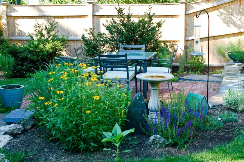 A backyard garden with blooming flowers, a birdbath, a metal bird feeder, and a patio set with four cushioned chairs on a brick patio, surrounded by greenery and enclosed by a wooden fence.