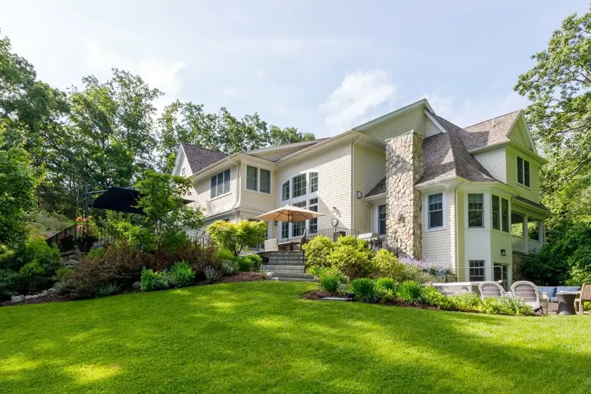 A large two-story white house with stone accents sits amid green lawns and lush landscaping, with outdoor seating and patio umbrellas visible on a sunny day.