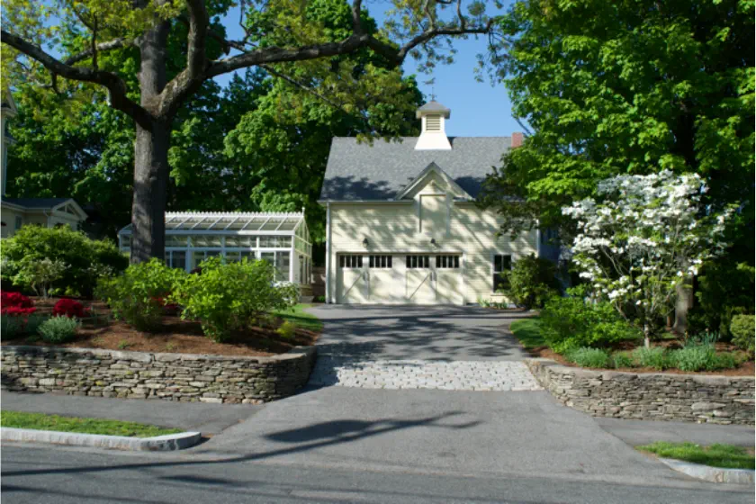 A charming light yellow house with a gray roof sits behind a stone retaining wall, surrounded by lush green trees and manicured gardens, with a glass greenhouse to the left.