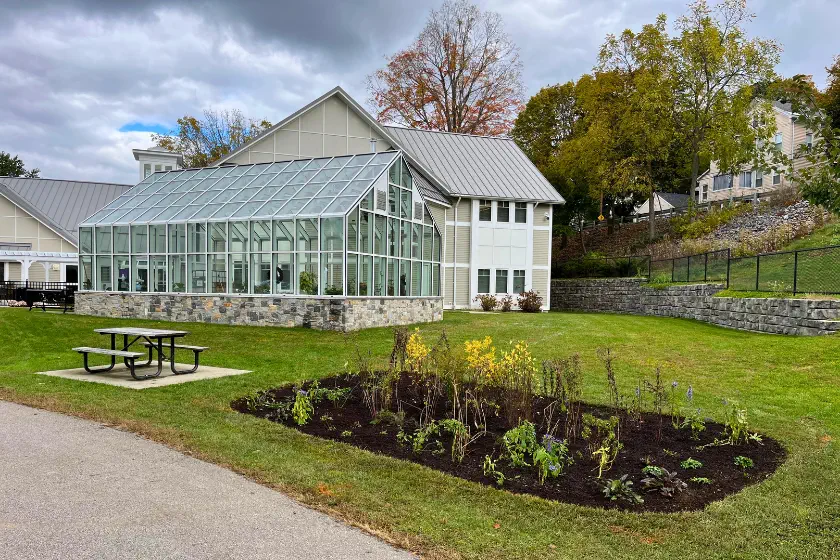 A modern greenhouse with many windows stands next to a white building. In the foreground, there is a small garden bed and a picnic table on a grassy lawn. Trees with autumn foliage are in the background under a cloudy sky.