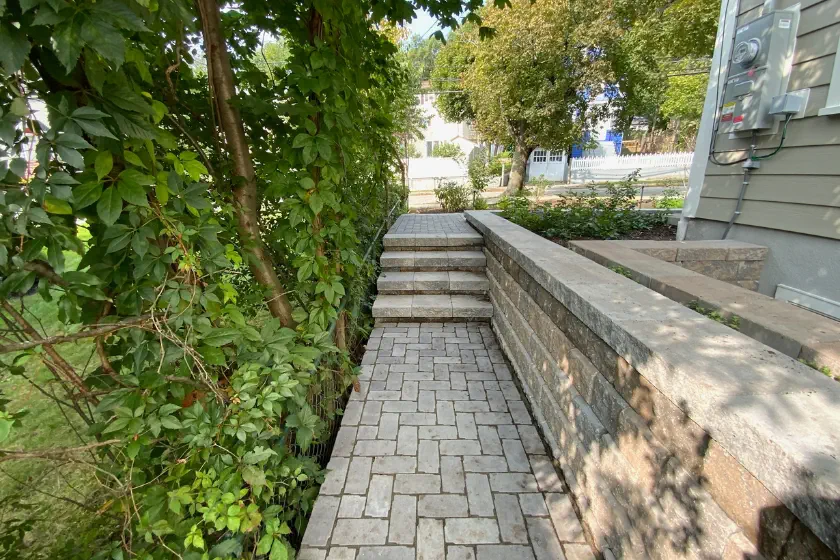 A paved brick walkway bordered by a stone wall and lush green vines leads up a short flight of steps toward a sunlit yard.