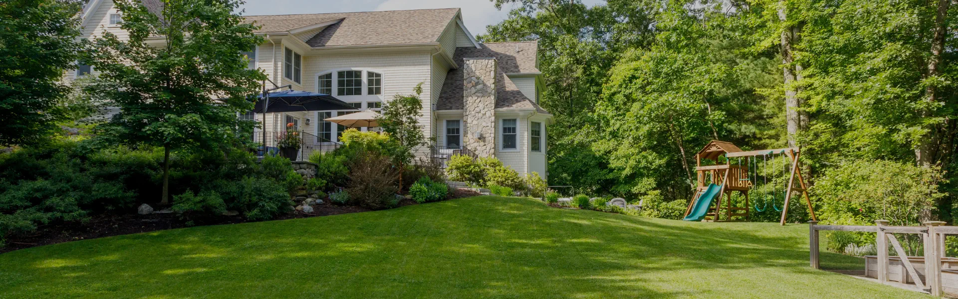 Large suburban house with stone accents, surrounded by lush green lawn, trees, a patio with umbrellas, a wooden playset with slide and swings, and a raised garden bed on a sunny day.