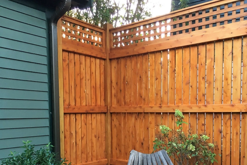 A wooden privacy fence with a decorative lattice top stands next to a green house, with some plants and patio furniture in the foreground.