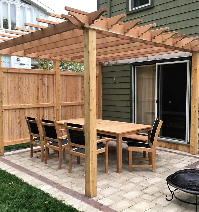 A wooden pergola shades a patio with a rectangular table and six chairs. The patio is paved with stone tiles, bordered by a wooden fence, and adjacent to a green house with sliding glass doors.