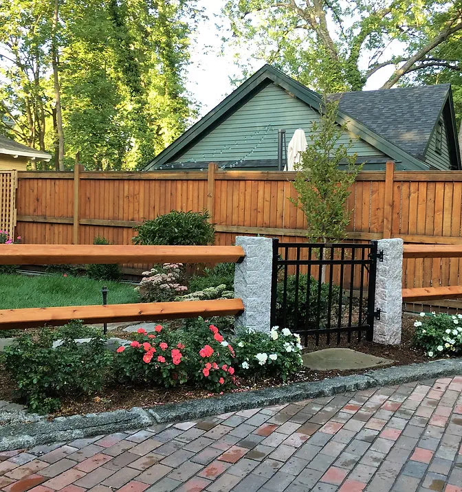 A backyard with a wooden fence, green lawn, flower beds with red and white roses, stone pillars, a black iron gate, and a small house with green siding in the background.