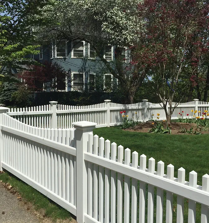 A white picket fence encloses a green yard with blooming trees, flowers, and a blue house in the background.