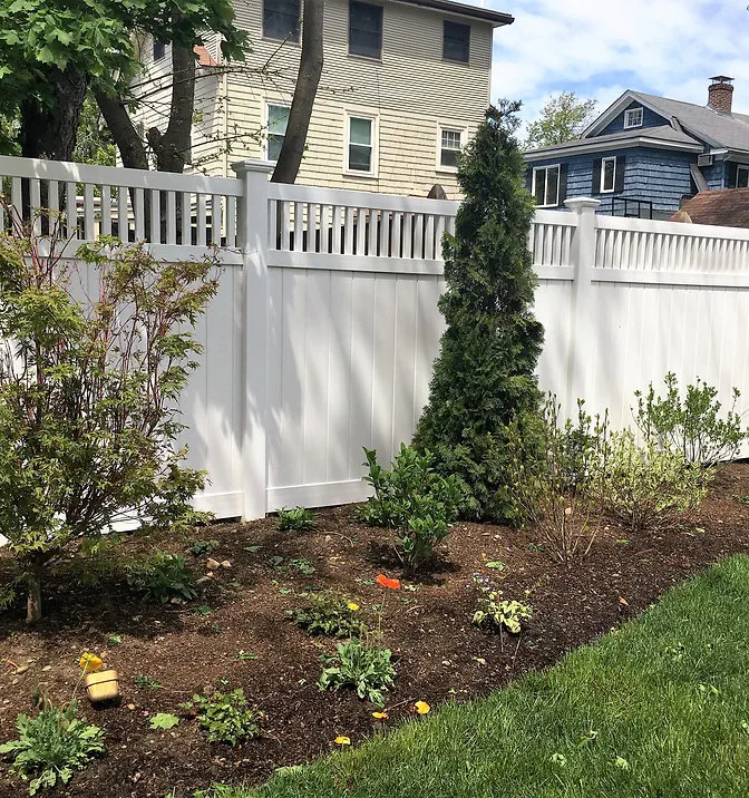 A garden bed with small plants and a few shrubs grows along a white fence, with green grass in the foreground and houses visible in the background.