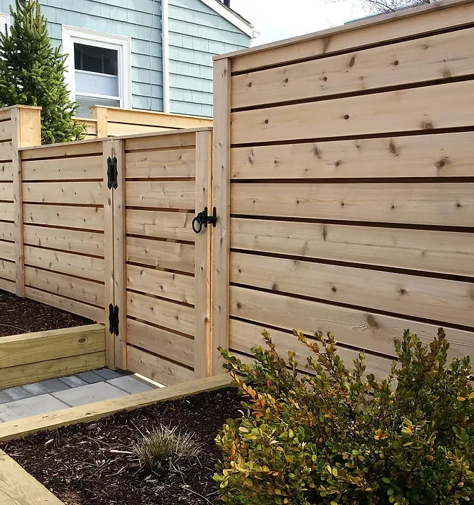 A modern wooden privacy fence with horizontal planks and a matching gate, accented by black metal hinges, surrounds a landscaped yard with shrubs and mulch near a house with blue siding.