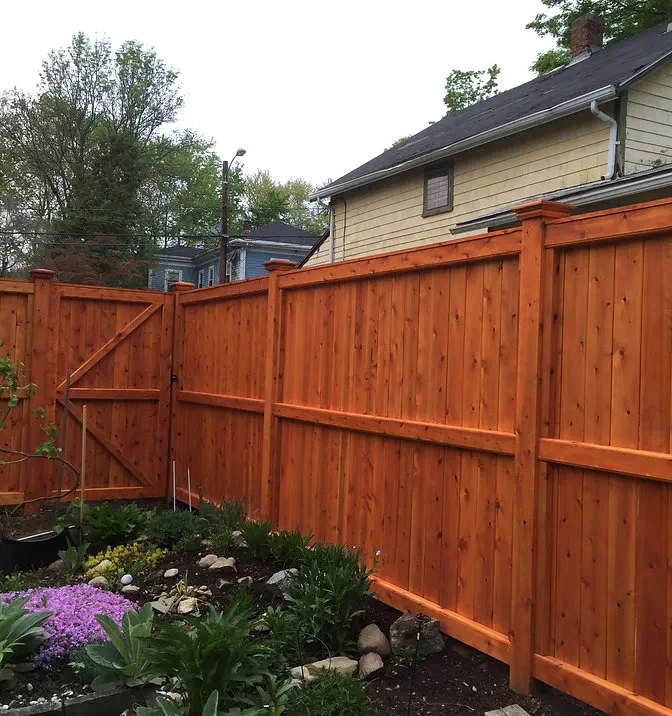 A backyard with a tall wooden privacy fence and gate, stained in a reddish-brown color. The fence surrounds a garden with green plants and purple flowers. Houses and trees are visible in the background.
