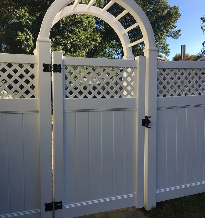 White vinyl fence with an arched gate featuring a lattice design at the top, black hinges, and surrounded by greenery and trees in the background.