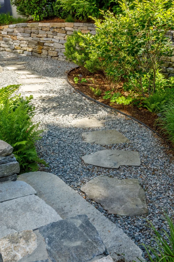 A garden path made of large flat stones set in gravel curves beside a raised stone wall and green leafy plants, creating a tranquil outdoor walkway.