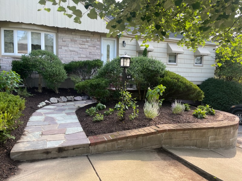 Garden Enhancement 1 A landscaped front yard with a stone walkway, curved brick edging, trimmed bushes, small plants, and a lantern near the steps offers beautiful garden enhancement to the entrance of a house with beige siding and stone facade.